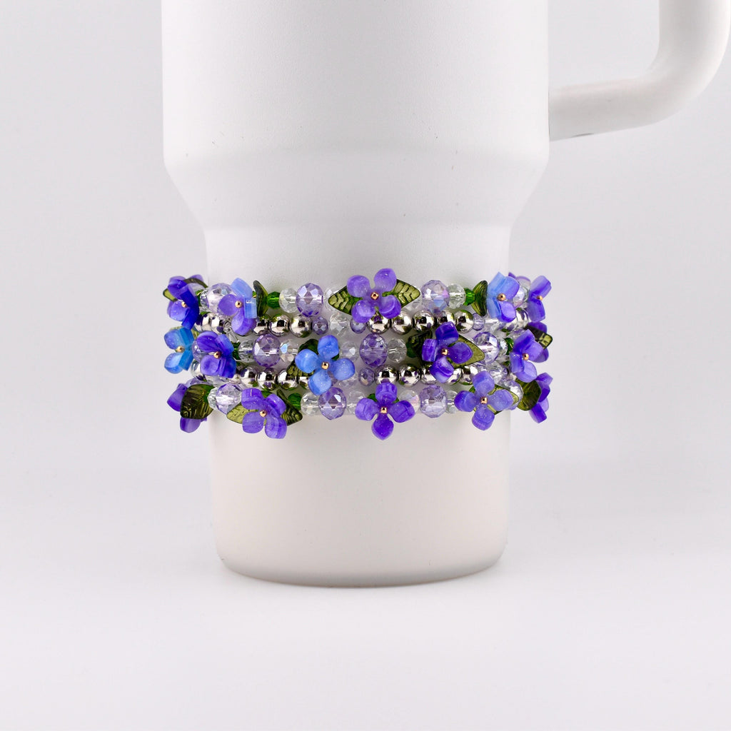 A Stanley tumbler is adorned with a bracelet featuring purple and blue flower beads. The bracelet is wrapped around the mug, showcasing the floral design and silver beads. The flowers have green leaves.