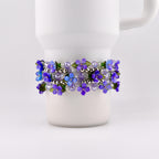 A Stanley tumbler is adorned with a bracelet featuring purple and blue flower beads. The bracelet is wrapped around the mug, showcasing the floral design and silver beads. The flowers have green leaves.