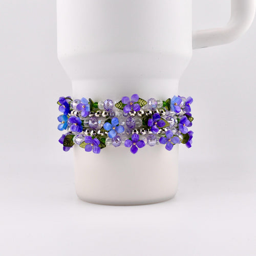 A Stanley tumbler is adorned with a bracelet featuring purple and blue flower beads. The bracelet is wrapped around the mug, showcasing the floral design and silver beads. The flowers have green leaves.