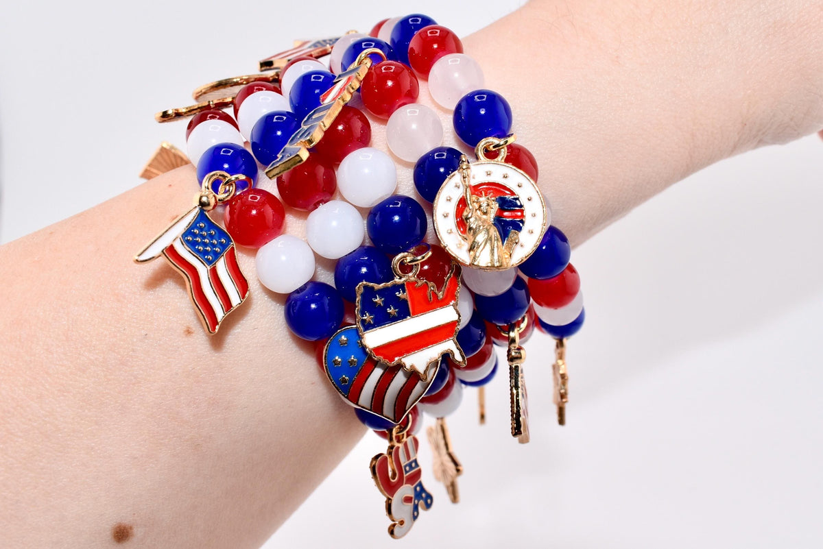 A patriotic bracelet featuring red, white, and blue beads with charms. Charms include an American flag, a map of the USA, and the Statue of Liberty. The bracelet is worn on a wrist.