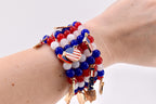 A beaded bracelet set featuring red, white, and blue beads. The bracelets are adorned with gold-colored charms, including American flags and patriotic symbols. The bracelet is worn on a wrist.