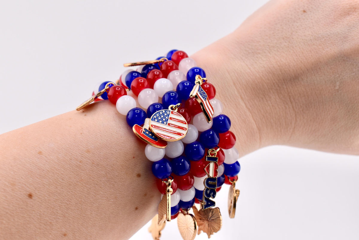 A beaded bracelet set featuring red, white, and blue beads. The bracelets are adorned with gold-colored charms, including American flags and patriotic symbols. The bracelet is worn on a wrist.
