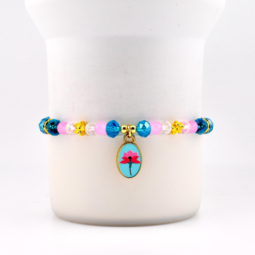 A beaded bracelet with a gold charm. The bracelet features pink, blue, yellow, and clear beads. The gold charm is an oval shape with a blue background and a pink flower design.