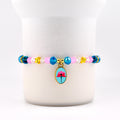 A beaded bracelet with a gold charm. The bracelet features pink, blue, yellow, and clear beads. The gold charm is an oval shape with a blue background and a pink flower design.