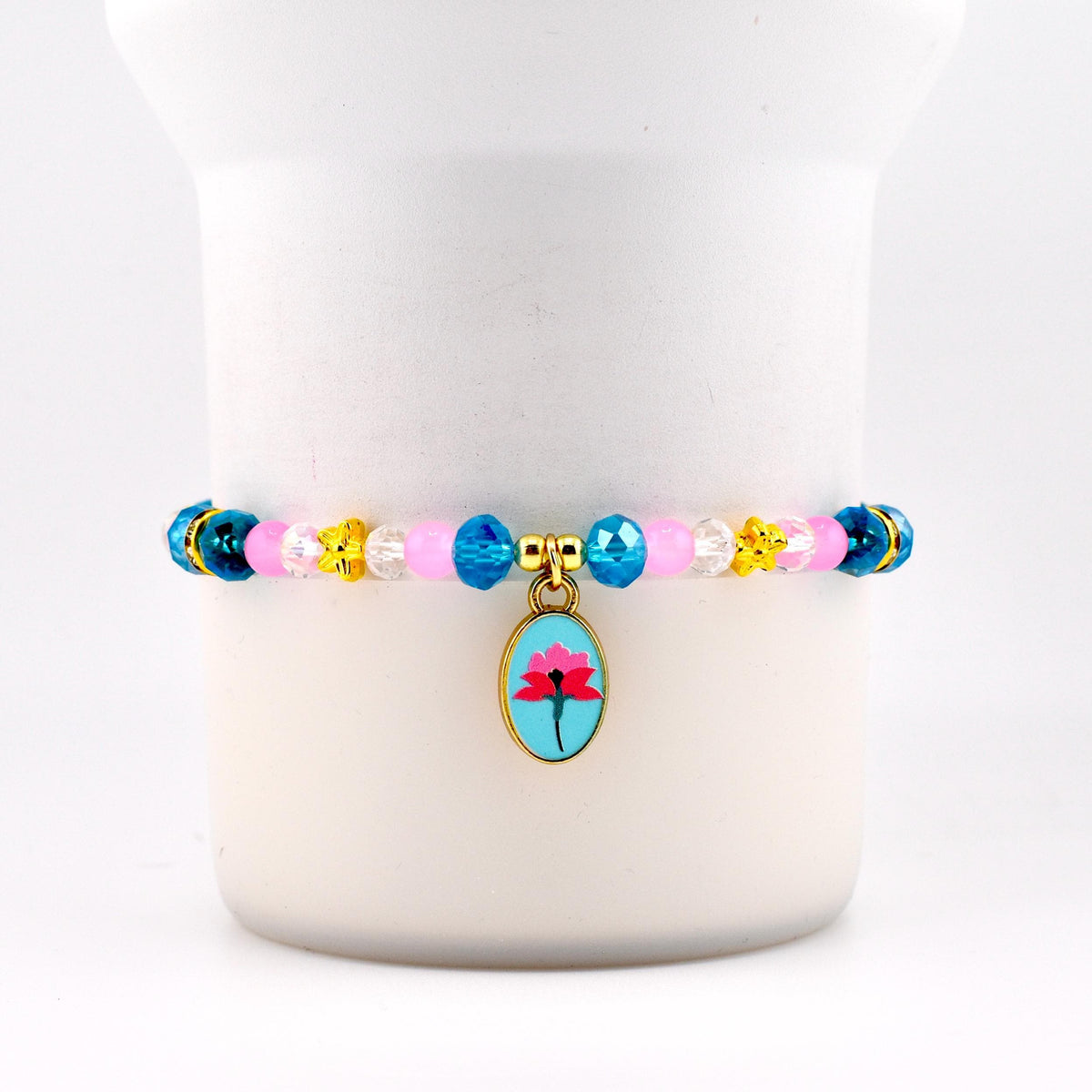 A beaded bracelet with a gold charm. The bracelet features pink, blue, yellow, and clear beads. The gold charm is an oval shape with a blue background and a pink flower design.