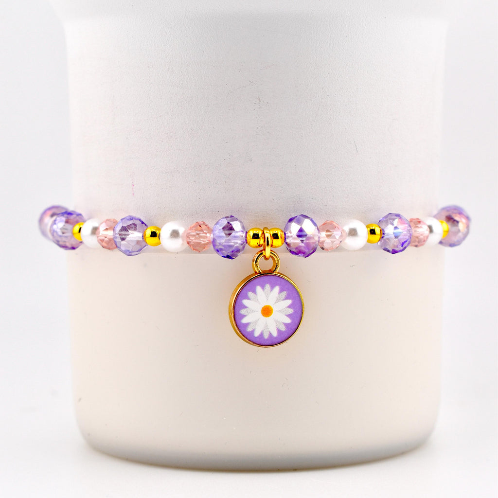 A beaded bracelet featuring a daisy charm. The bracelet has purple, pink, and white beads with gold accents. The daisy charm is purple with a white flower design.