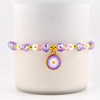 A beaded bracelet featuring a daisy charm. The bracelet has purple, pink, and white beads with gold accents. The daisy charm is purple with a white flower design.