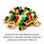 A stack of beaded bracelets with red, yellow, green, and black beads. The bracelets have gold-colored charms with Juneteenth themes, including flags and the text "SINCE 1865".