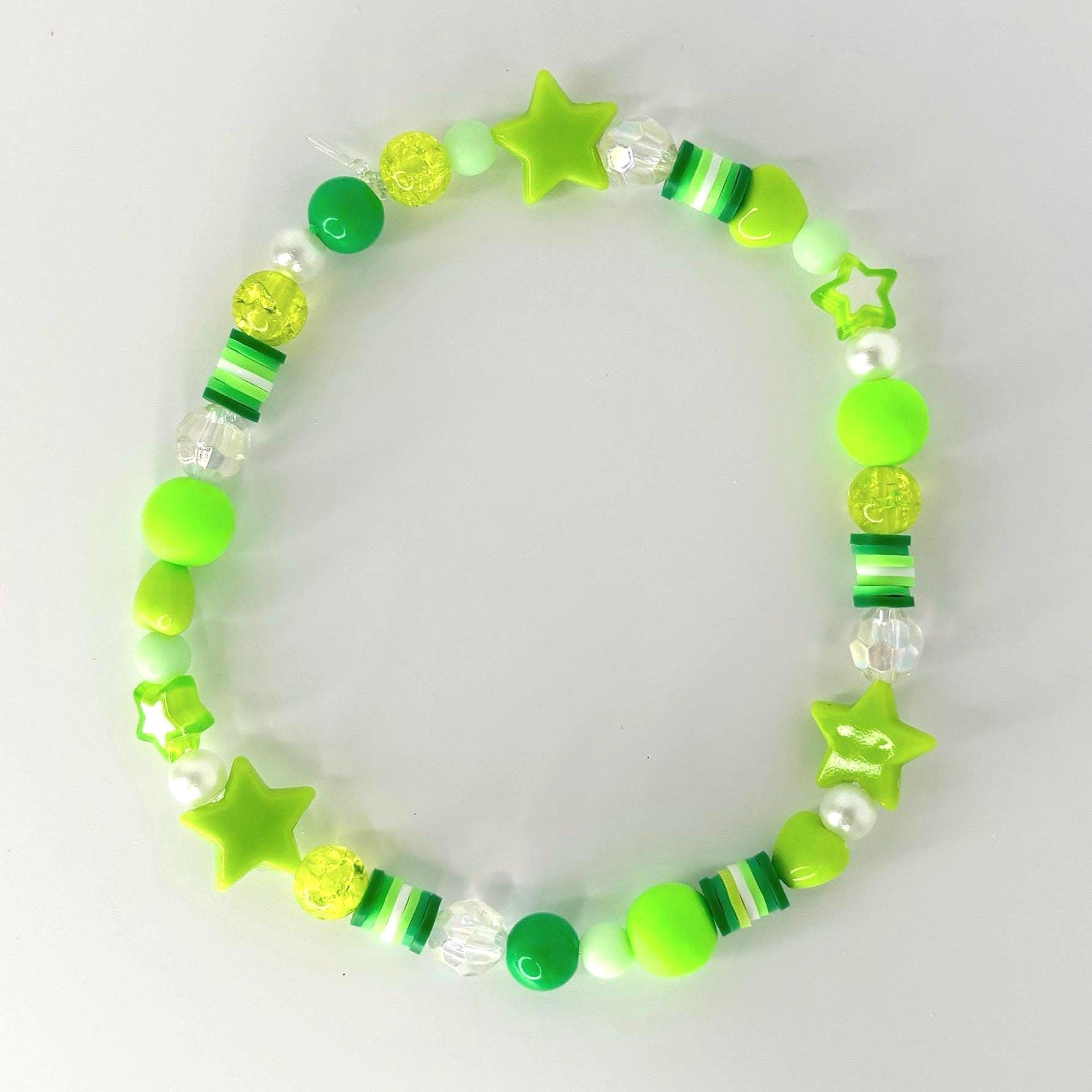 A beaded bracelet featuring a variety of green beads, including round, star-shaped, and cylindrical beads. The bracelet incorporates shades of lime, neon, and forest green, with clear and white accents. The bracelet is designed with a playful and youthful aesthetic.