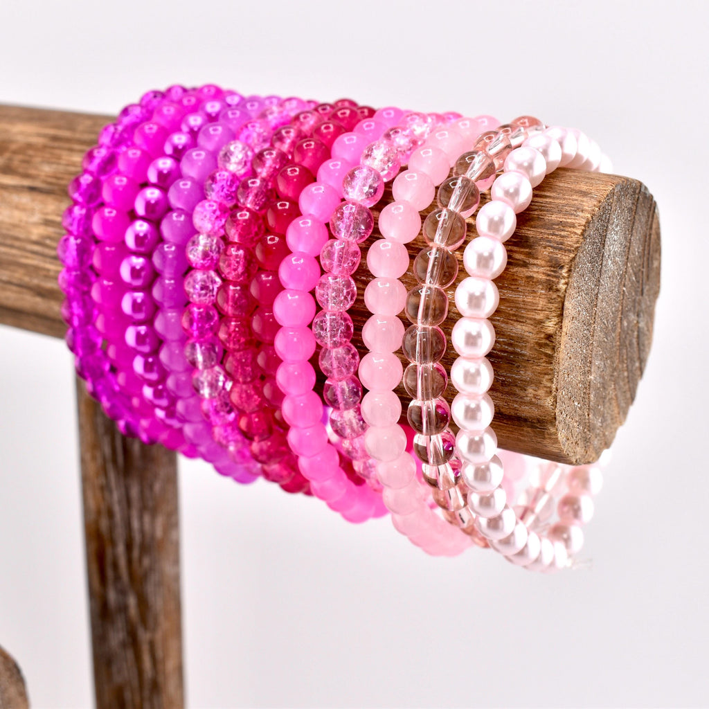 A collection of pink beaded bracelets in various shades, from deep magenta to pale pink, displayed on a wooden stand. The bracelets are made of small, round beads and are arranged in a gradient of color.