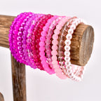 A collection of pink beaded bracelets in various shades, from deep magenta to pale pink, displayed on a wooden stand. The bracelets are made of small, round beads and are arranged in a gradient of color.