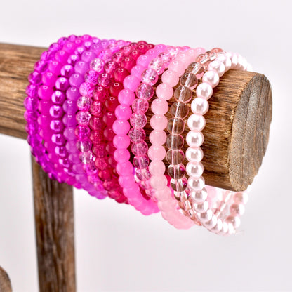 A collection of pink beaded bracelets in various shades, from deep magenta to pale pink, displayed on a wooden stand. The bracelets are made of small, round beads and are arranged in a gradient of color.