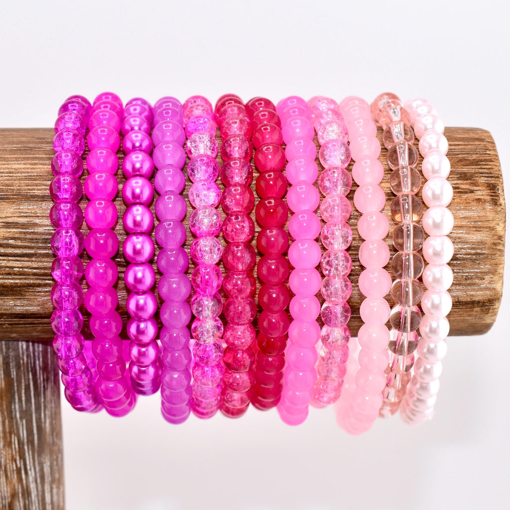 A collection of pink beaded bracelets in various shades, from deep magenta to pale blush, displayed on a wooden stand. The bracelets feature different bead styles, including smooth, faceted, and pearl-like beads, creating a diverse and colorful accessory set.