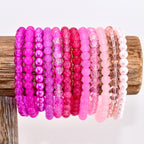 A collection of pink beaded bracelets in various shades, from deep magenta to pale blush, displayed on a wooden stand. The bracelets feature different bead styles, including smooth, faceted, and pearl-like beads, creating a diverse and colorful accessory set.