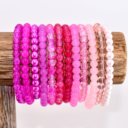 A collection of pink beaded bracelets in various shades, from deep magenta to pale blush, displayed on a wooden stand. The bracelets feature different bead styles, including smooth, faceted, and pearl-like beads, creating a diverse and colorful accessory set.