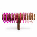 A wooden bracelet display stand holding a collection of beaded bracelets in shades of pink and purple. The bracelets are arranged in a gradient of colors, from deep magenta to pale pink. The stand is made of dark wood.