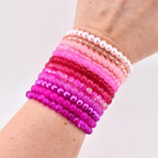 A collection of pink bracelets in various shades, from light pink to deep magenta, are stacked on a wrist. The bracelets include pearl, beaded, and clear styles, creating a gradient effect. The bracelets are a fashion accessory.