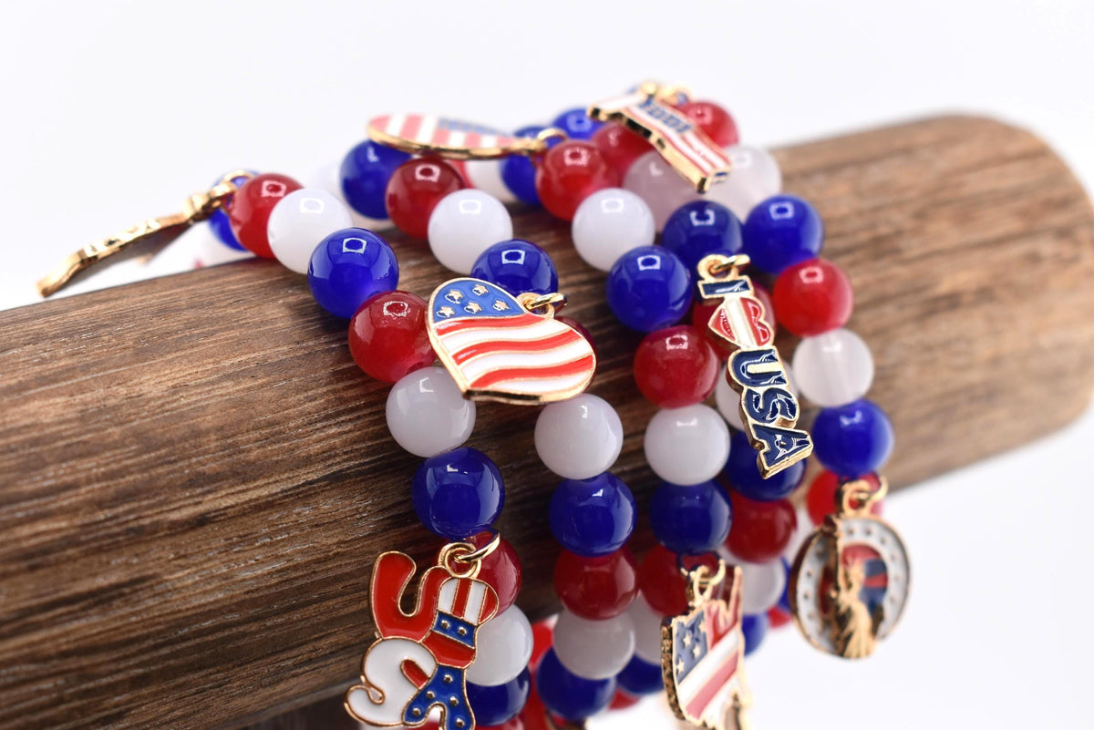 Beaded bracelets in red, white, and blue, adorned with patriotic charms. The bracelets feature round beads and charms shaped like the American flag, a top hat, and a star. The bracelets are displayed on a wooden surface.