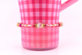 A pink beaded bracelet with gold accents and a central clear bead is displayed against a pink gingham background. The bracelet features various shades of pink beads and gold spacers. The background is a pink gingham pattern on a mug.