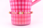 A pink beaded bracelet with gold accents and a central clear bead is displayed against a pink gingham background. The bracelet features various shades of pink beads and gold spacers. The background is a pink gingham pattern on a mug.
