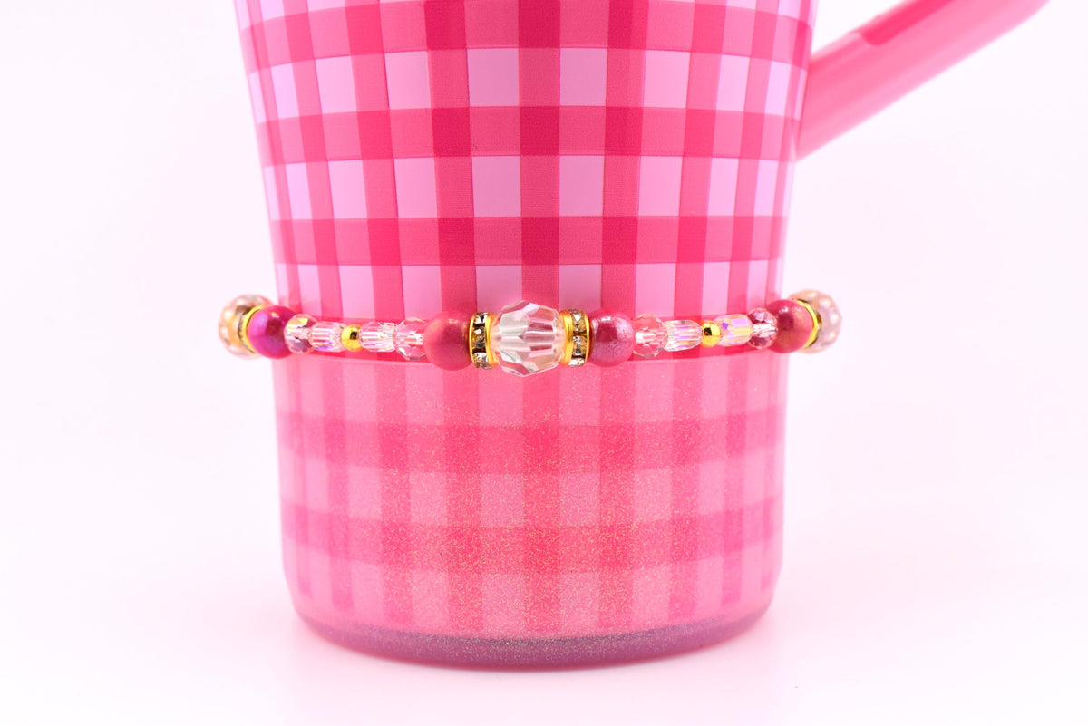 A pink beaded bracelet with gold accents and a central clear bead is displayed against a pink gingham background. The bracelet features various shades of pink beads and gold spacers. The background is a pink gingham pattern on a mug.