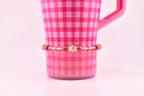 A pink and gold beaded bracelet with a central clear bead and smaller pink and gold beads. The bracelet is displayed in front of a pink checkered mug.