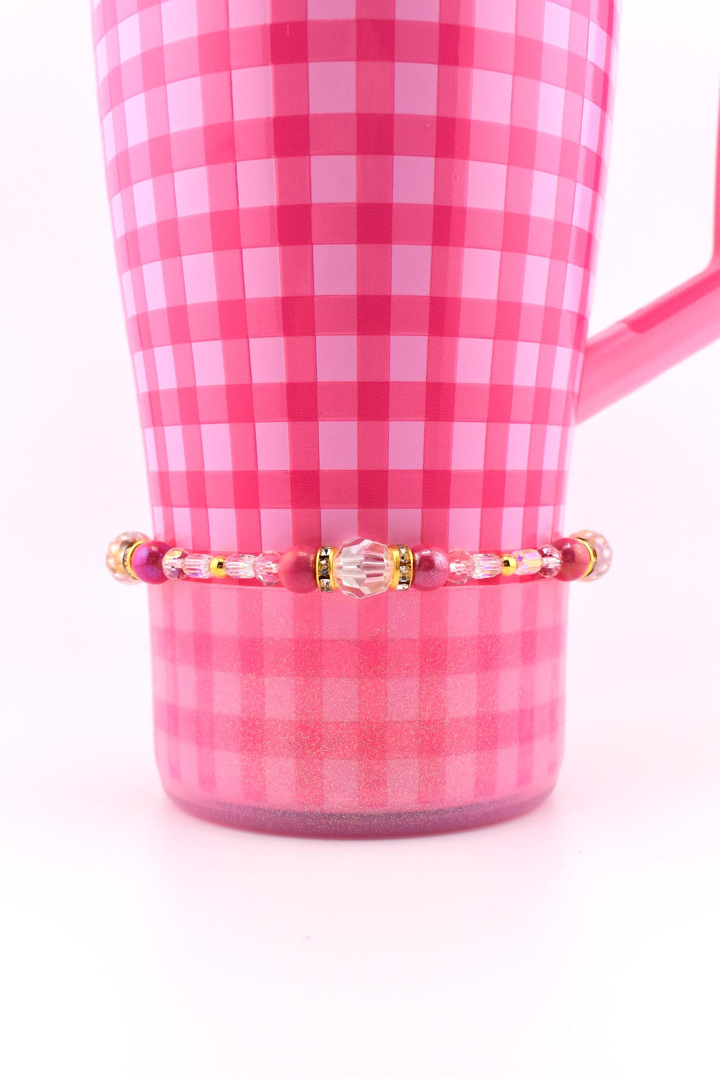 A pink and white checkered mug with a beaded bracelet. The bracelet features a variety of pink and clear beads, with gold accents. The mug has a handle and is set against a white background.