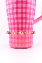 A pink and white checkered mug with a beaded bracelet. The bracelet features a variety of pink and clear beads, with gold accents. The mug has a handle and is set against a white background.