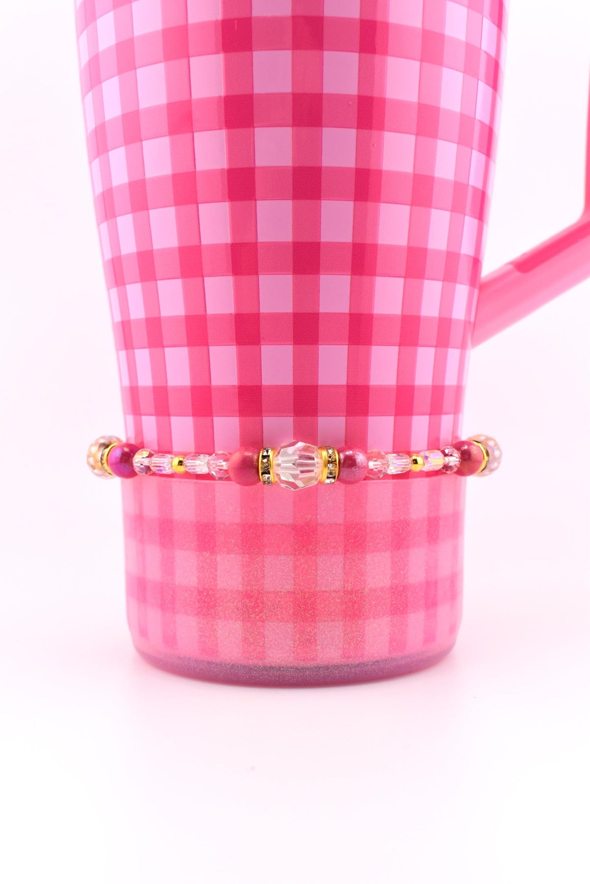 A pink and white checkered mug with a beaded bracelet. The bracelet features a variety of pink and clear beads, with gold accents. The mug has a handle and is set against a white background.