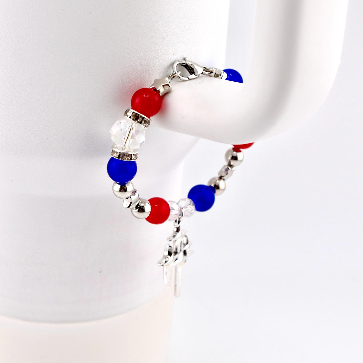 A patriotic bracelet featuring red, white, and blue beads, interspersed with silver-colored beads. A small silver charm hangs from the bracelet. The bracelet is secured with a silver clasp.