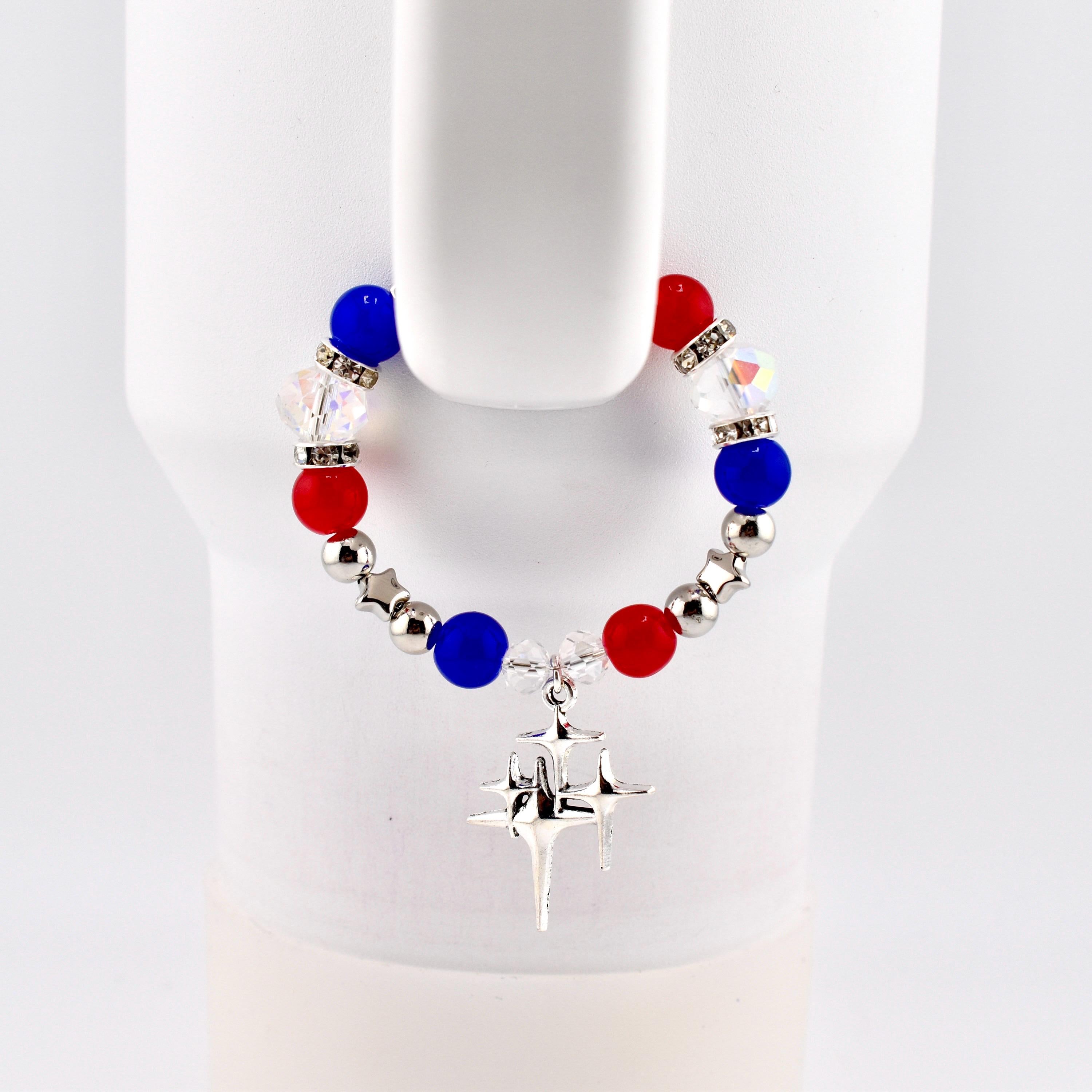 A patriotic bracelet featuring red, white, and blue beads, interspersed with silver-colored beads and star-shaped charms. The bracelet is adorned with a dangling silver star pendant. The beads are approximately 0.5 inches in diameter.