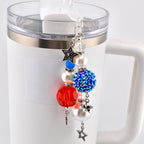 A beaded charm with red, white, and blue beads and star charms hangs from a white mug. The charm features a faceted red bead, a blue rhinestone-covered bead, and white pearl-like beads.