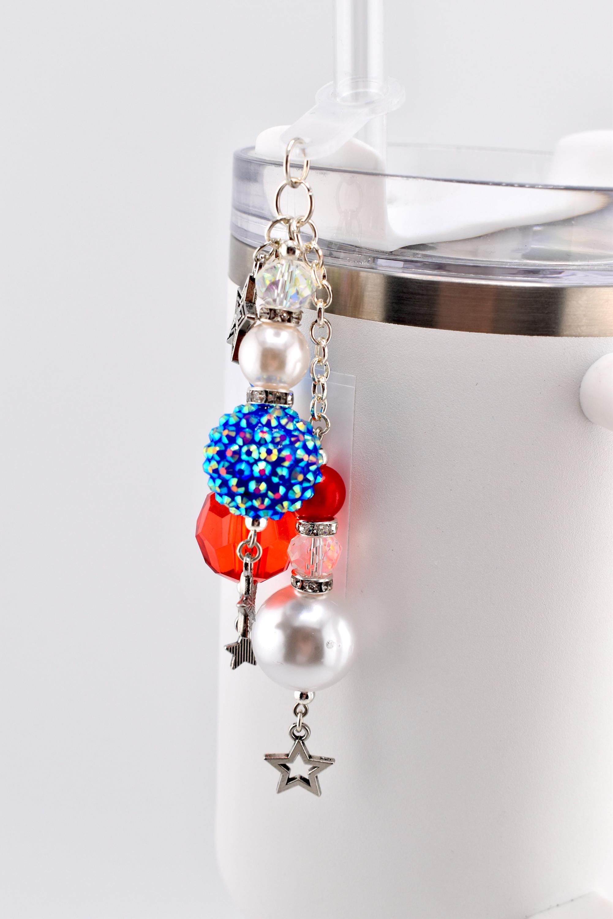 A beaded charm in red, white, and blue hues, attached to a silver chain. The charm features a mix of beads, including a large blue rhinestone-covered ball, red faceted beads, and white pearl-like beads. Silver star charms dangle from the bottom.