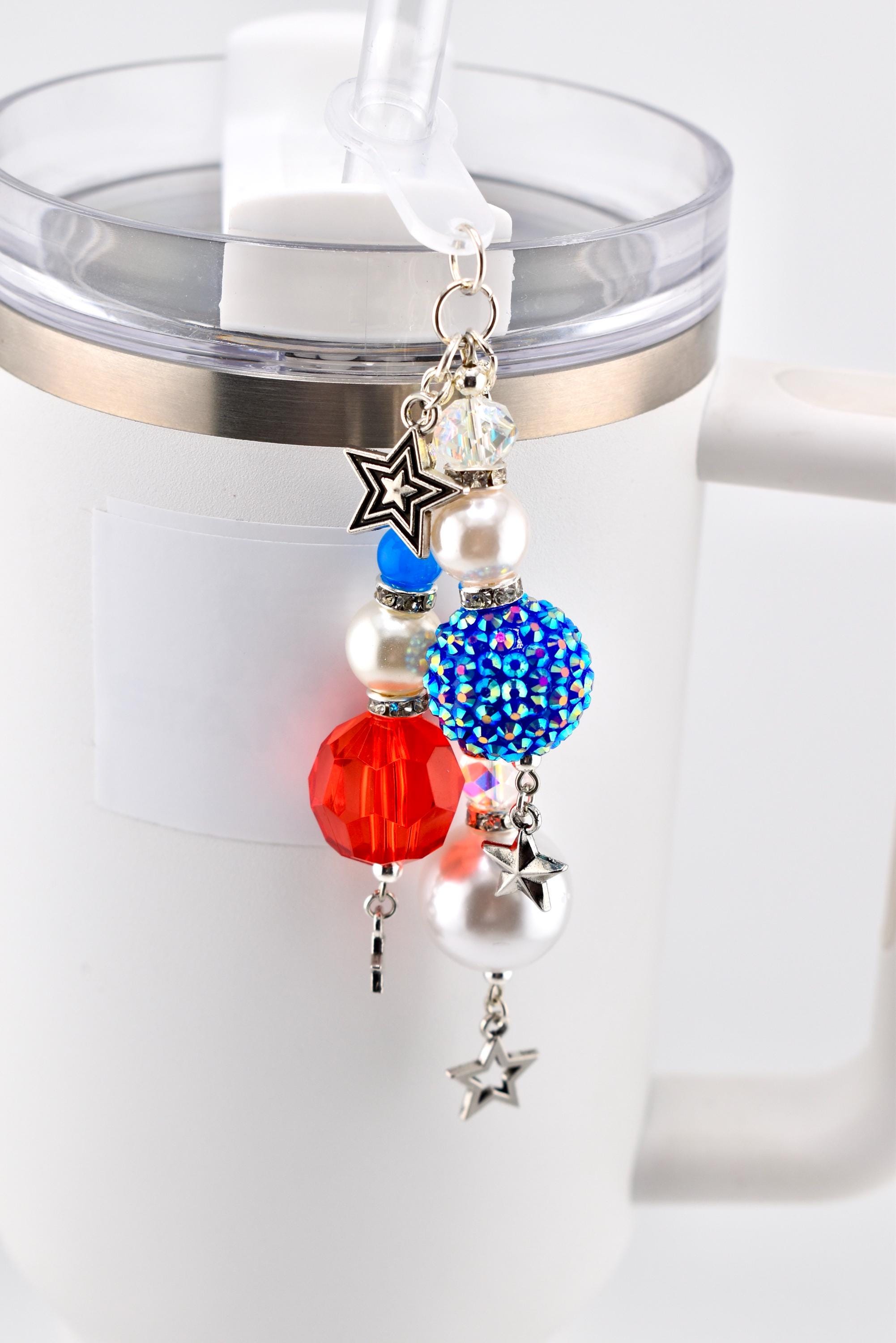 A patriotic-themed charm with red, white, and blue beads and star accents. The charm is attached to a white tumbler with a silver rim and a clear straw. The charm includes a faceted red bead, a blue rhinestone-covered bead, and pearl beads.