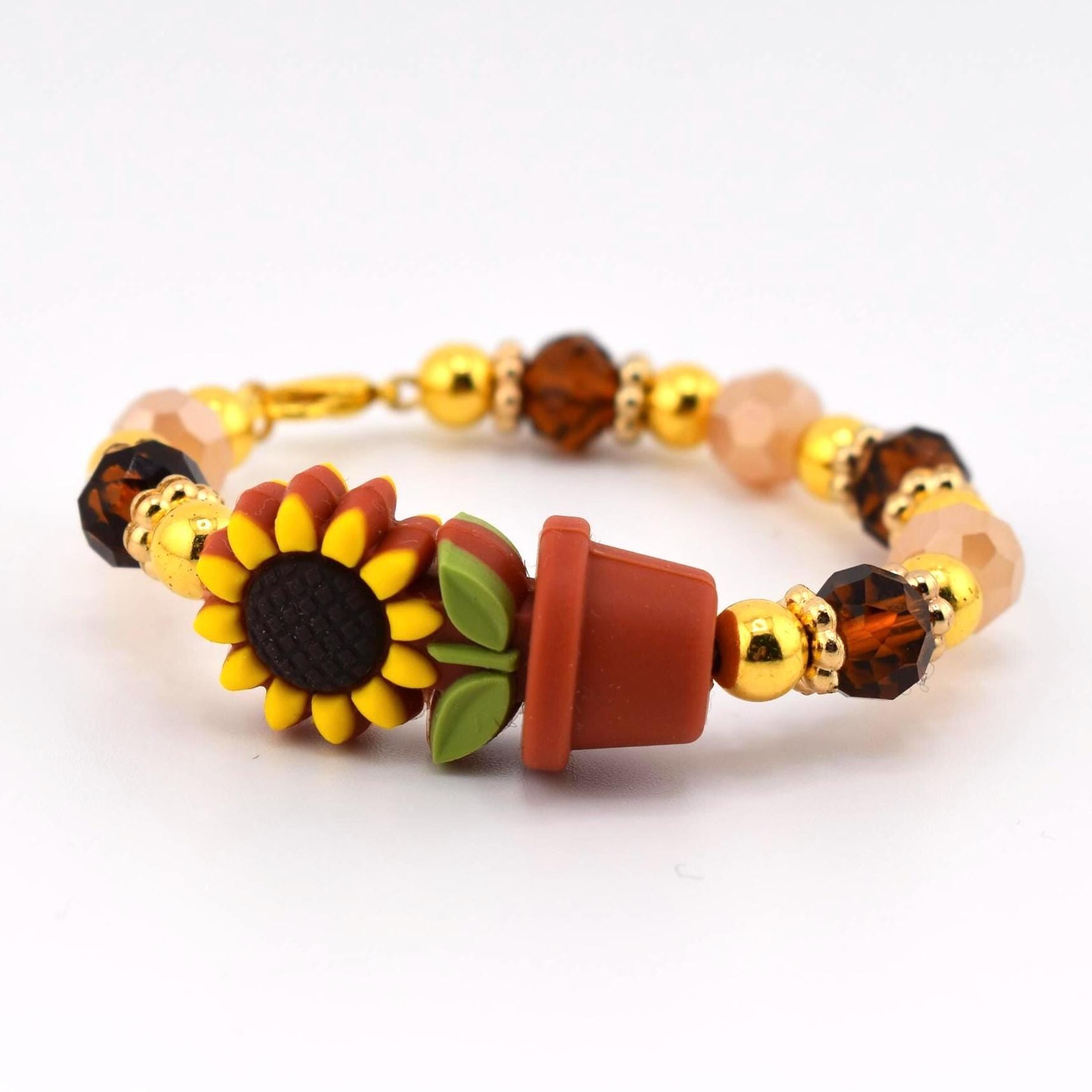 A beaded bracelet featuring a sunflower and a flower pot charm. The bracelet has gold-colored beads, brown and peach-colored faceted beads, and a gold-colored clasp. The sunflower has yellow petals and a dark center.