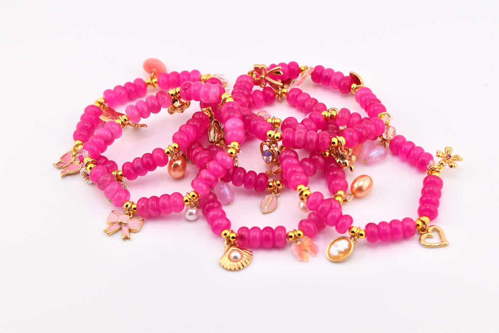 A collection of vibrant pink beaded bracelets with gold-colored charms. The bracelets feature various charms, including bows, hearts, and seashell pendants. The beads are a deep magenta color, and the bracelets are arranged on a white surface.