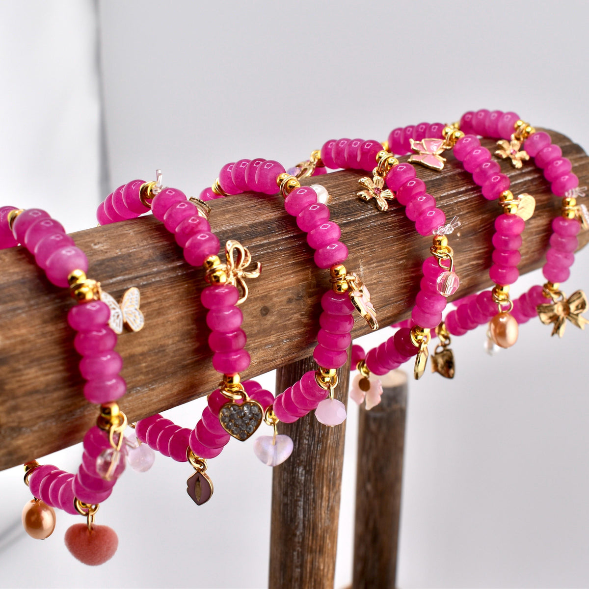 A collection of pink beaded bracelets with gold-colored charms, including butterflies, bows, and hearts. The bracelets are displayed on a wooden stand, showcasing their vibrant color and intricate details. Jewelry.