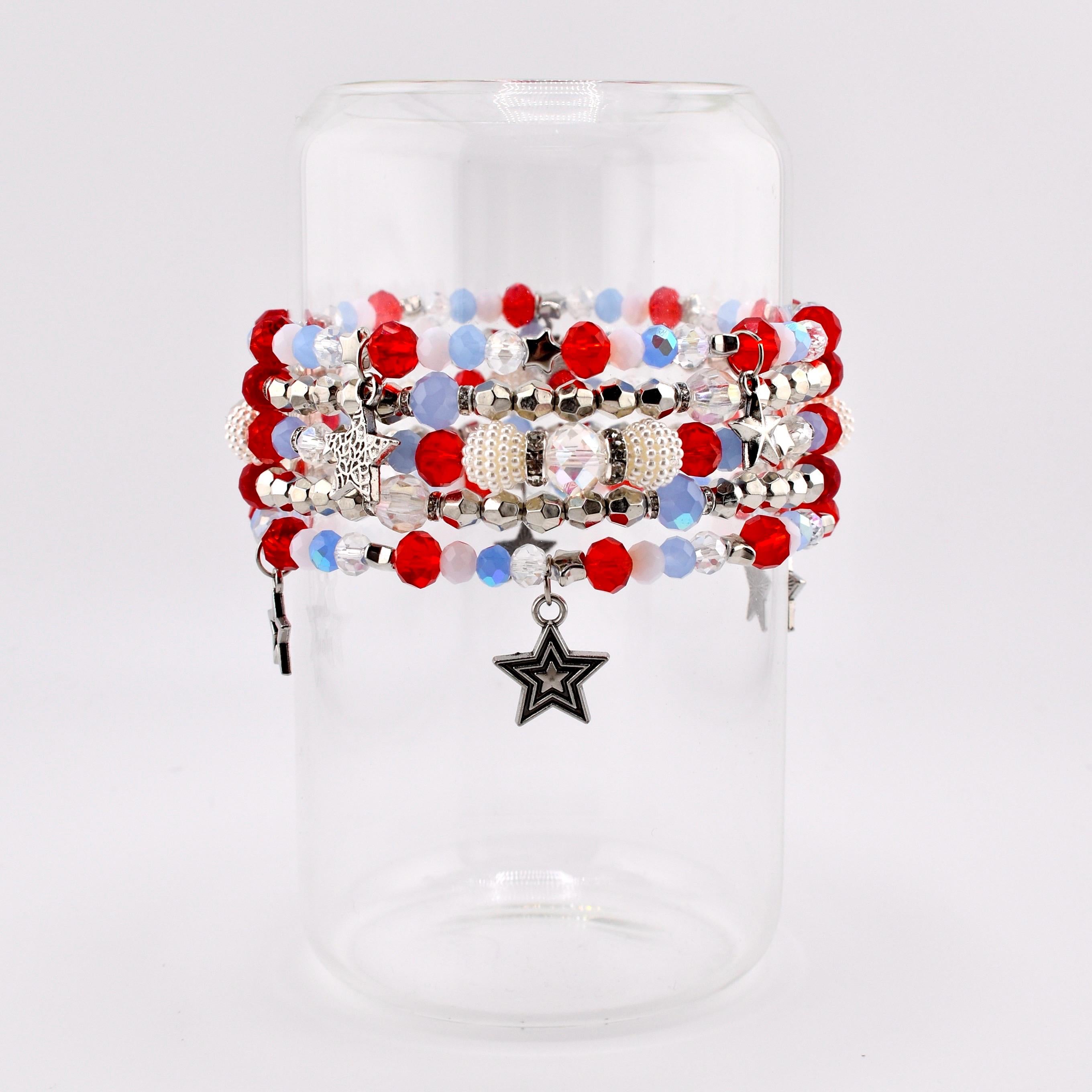 A collection of beaded bracelets in red, white, and blue, with silver accents and star charms. The bracelets are displayed on a clear glass cylinder, showcasing the patriotic color scheme and intricate beadwork. The bracelets feature a variety of bead sizes and textures.