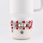 A stack of beaded bracelets in red, white, and blue, with star charms. The bracelets feature various bead sizes and textures, including small red beads, iridescent beads, and white pearl-like beads. The bracelets are stacked on a white mug.