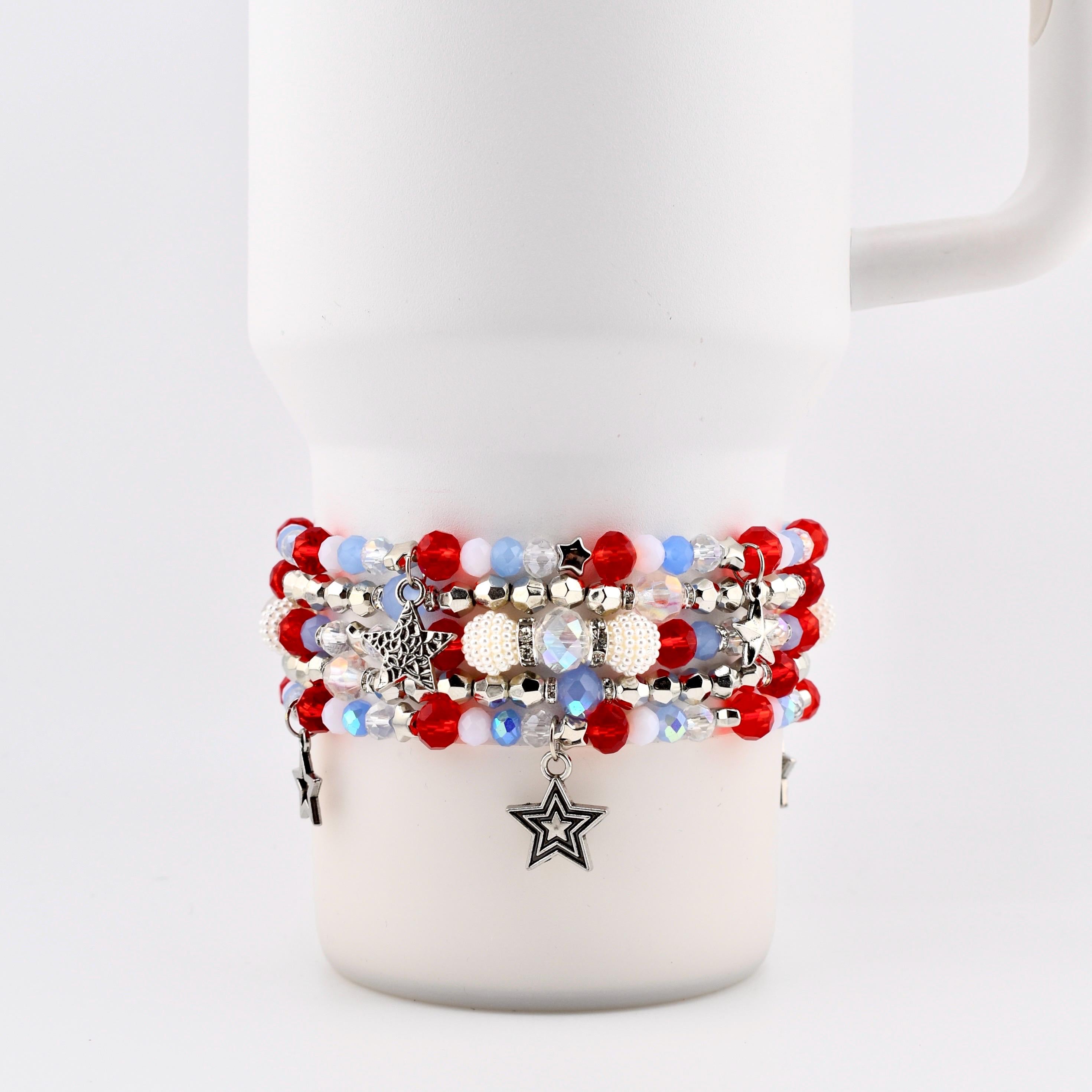 A stack of beaded bracelets in red, white, and blue, with star charms. The bracelets feature various bead sizes and textures, including small red beads, iridescent beads, and white pearl-like beads. The bracelets are stacked on a white mug.