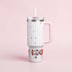 A white tumbler with pink stripes and star accents in red, white, and blue. The tumbler has a pink handle, lid, and straw. A beaded bracelet with star charms adorns the base of the tumbler.