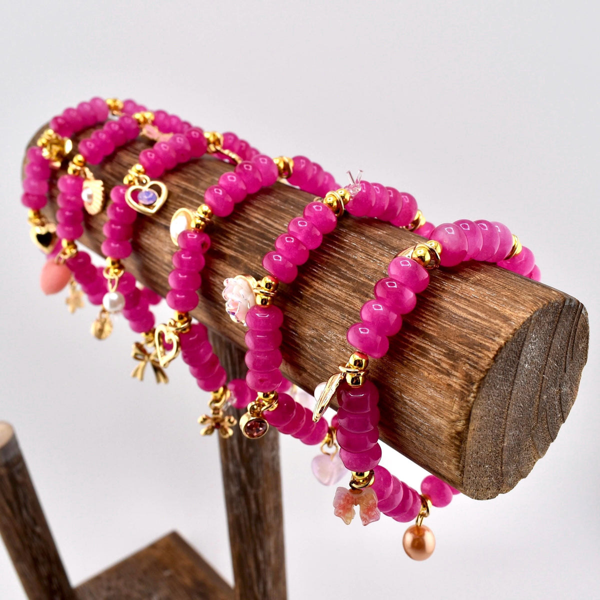 Several fuchsia beaded bracelets with gold-colored charms and accents are displayed on a wooden stand. The charms include hearts, bows, and other decorative elements. The bracelets are arranged in a row, showcasing their design.
