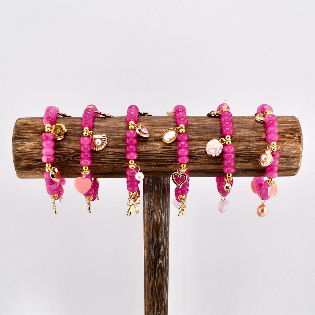 Six pink beaded bracelets with gold accents and charms, including hearts, shells, and bows, are displayed on a wooden stand. The bracelets are made of small, round beads and are designed for wrist wear.