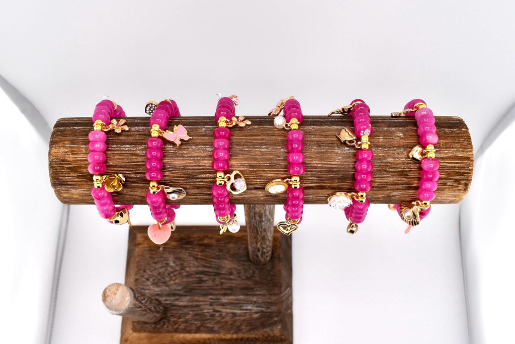 Six pink beaded bracelets with gold-colored charms, including hearts, butterflies, and other shapes, are displayed on a wooden stand. The bracelets are made of small, round beads and are arranged in a row. The charms add a decorative touch.