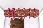 Six pink beaded bracelets with gold-colored charms, including hearts, butterflies, and other shapes, are displayed on a wooden stand. The bracelets are made of small, round beads and are arranged in a row. The charms add a decorative touch.