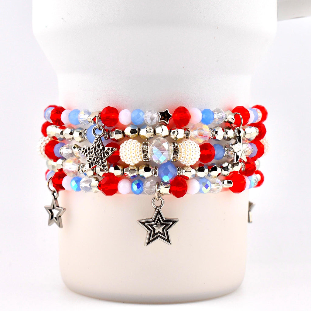 A stack of beaded bracelets in red, white, and blue. The bracelets feature various bead sizes and textures, with silver star charms dangling from the bottom. Some beads are iridescent, and others are pearl-like.