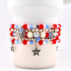 A stack of beaded bracelets in red, white, and blue. The bracelets feature various bead sizes and textures, with silver star charms dangling from the bottom. Some beads are iridescent, and others are pearl-like.