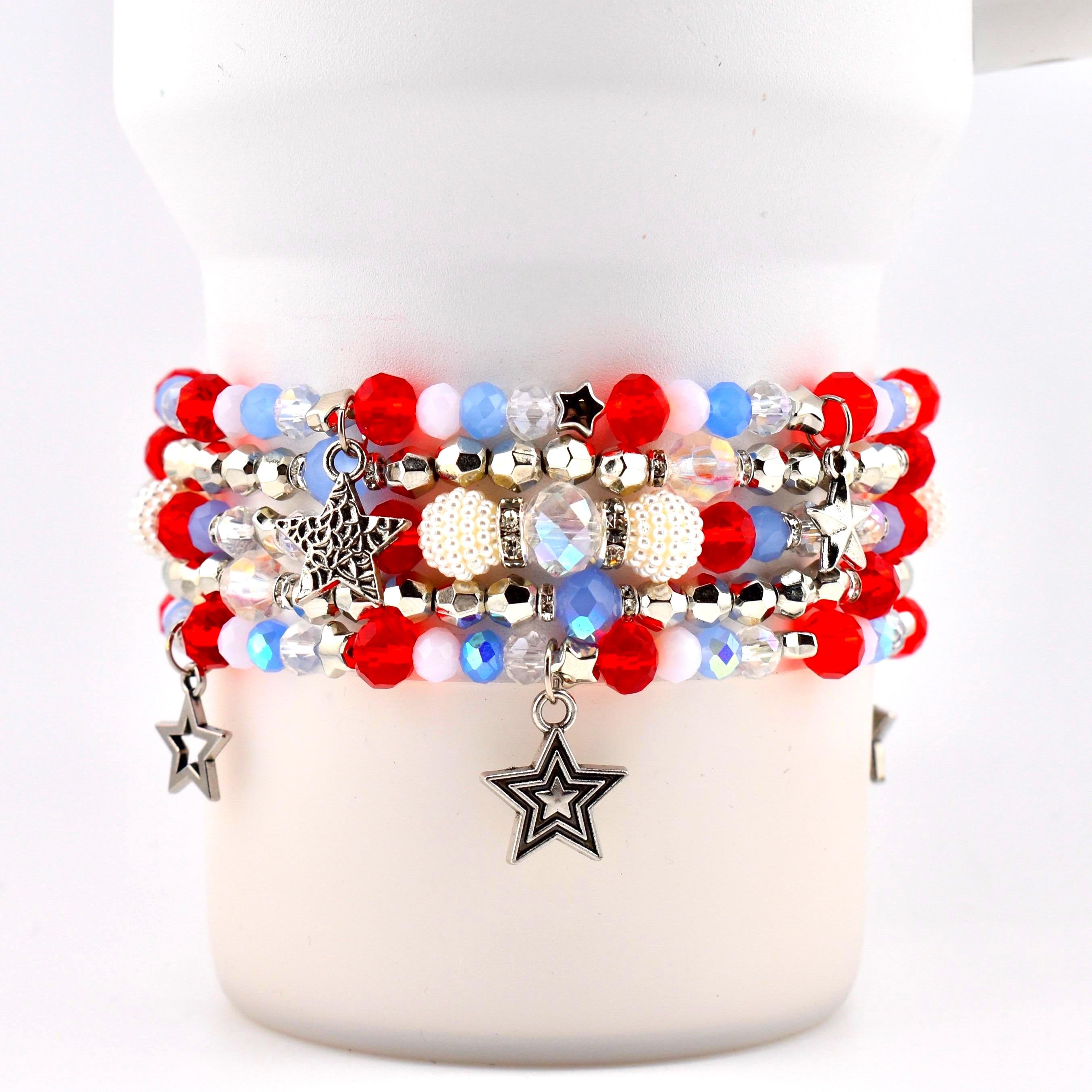 A stack of beaded bracelets in red, white, and blue. The bracelets feature various bead sizes and textures, with silver star charms dangling from the bottom. Some beads are iridescent, and others are pearl-like.