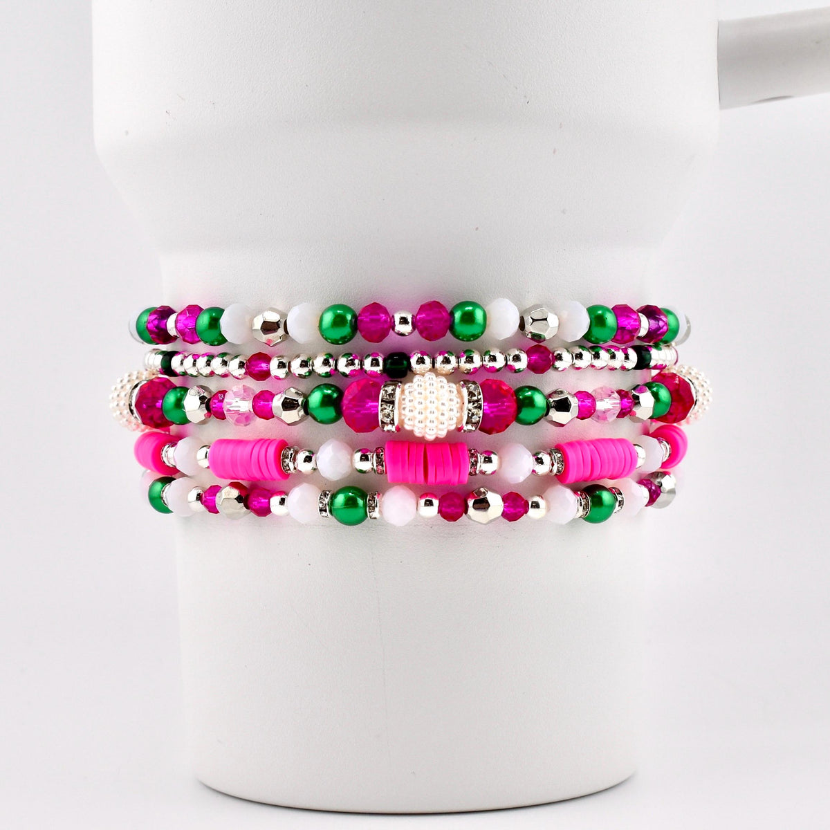 A stack of five beaded bracelets in shades of pink, green, white, and silver. The bracelets feature various bead sizes and textures, including small round beads, larger faceted beads, and flat disc beads. The bracelets are displayed on a white background.