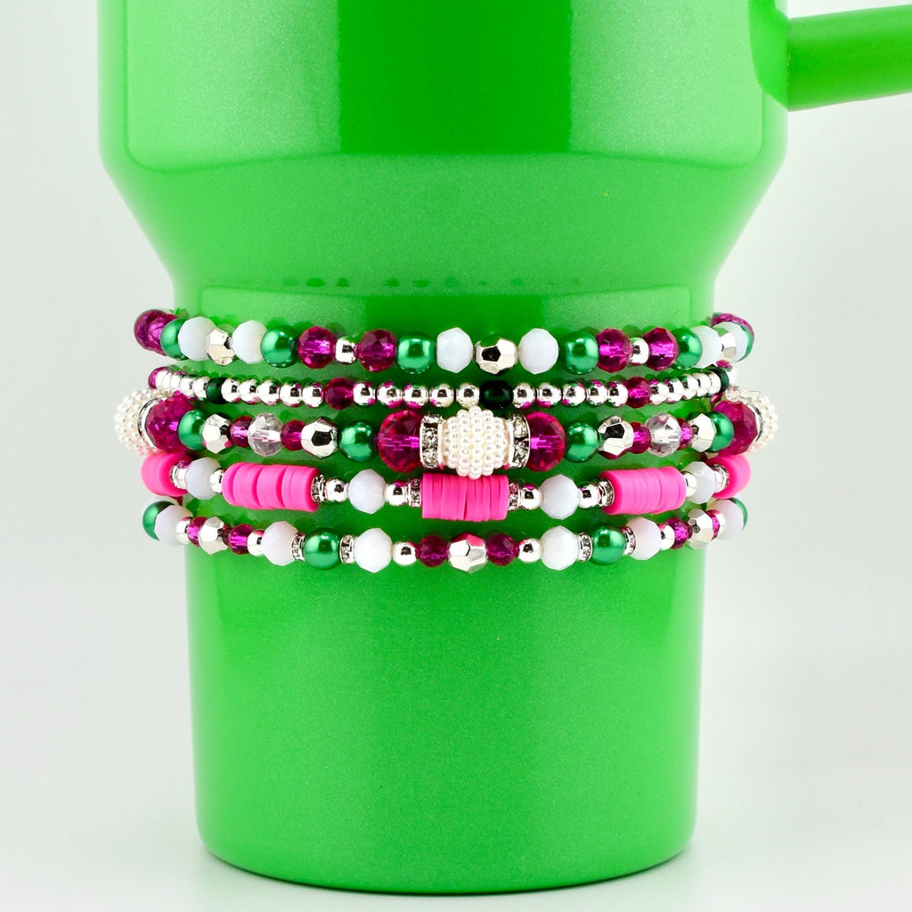 A collection of beaded bracelets in shades of pink, green, white, and silver. The bracelets are stacked on a bright green tumbler. The beads vary in size and material, creating a layered and textured look. The bracelets are a mix of round and flat beads.